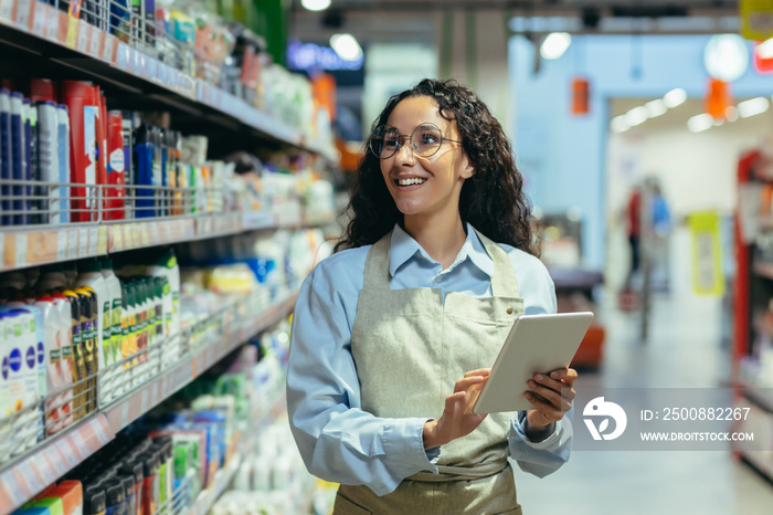 A young Hispanic woman works in a supermarket. Stands in uniform in the department of household chemicals with a tablet, conducts an audit, makes a list, checks the goods.