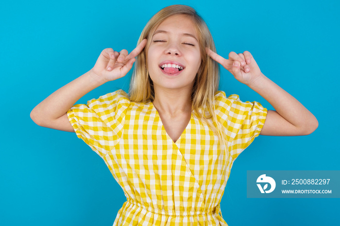 Photo of crazy beautiful Caucasian little girl wearing yellow dress over blue background screaming and pointing with fingers at hair closed eyes