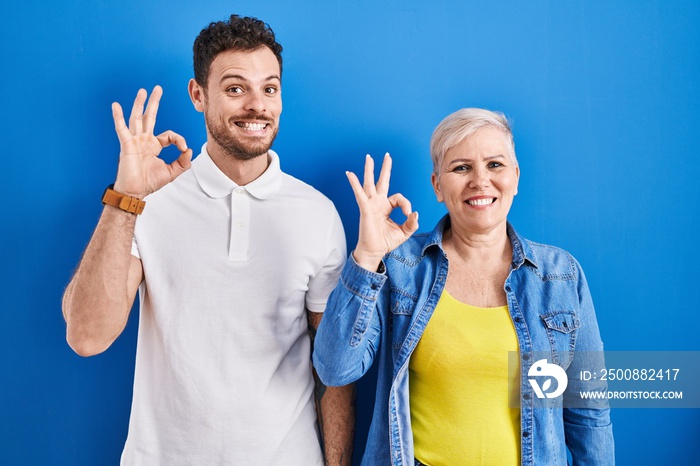 Young brazilian mother and son standing over blue background smiling positive doing ok sign with hand and fingers. successful expression.