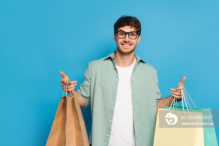 happy young man looking at camera while holding shopping bags on blue