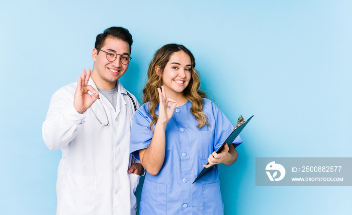 Young doctor couple posing in a blue background isolated cheerful and confident showing ok gesture.