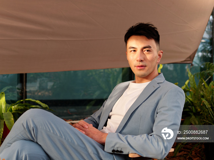 Portrait of handsome Chinese young man in light blue suit and white undershirt sitting and looking away with legs crossed on knee in sunny day, front view of confident businessman.