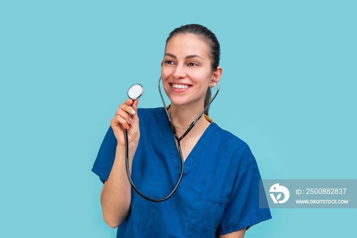 Studio portrait of a happy and smiling brunette doctor woman or nurse wearing blue scrubs uniform holding stethoscope, isolated on blue background