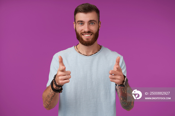 Happy young attractive brunette man with beard looking at camera cheerfully and smiling, raising hands with thumbs and showing his pleasant emotions, isolated over purple background