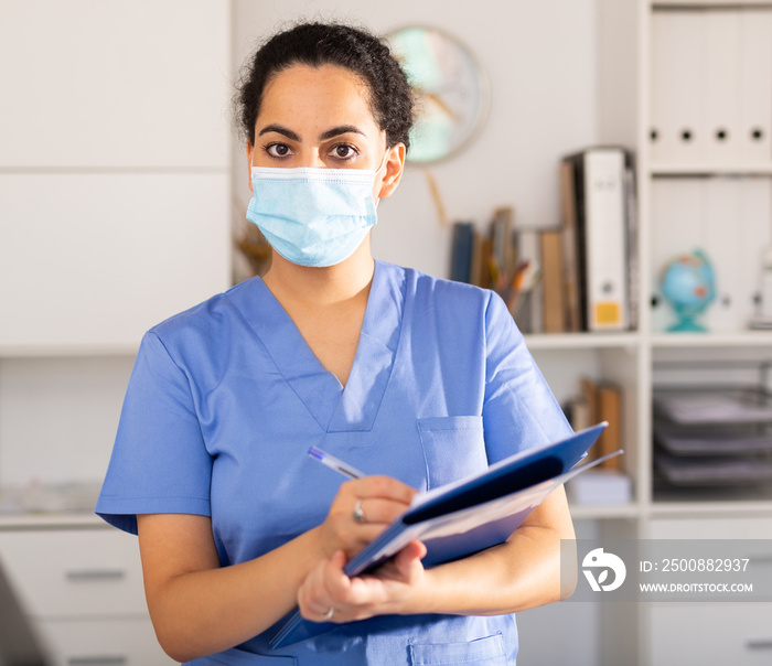 Positive female doctor wearing face mask and blue overall meeting client before consultation