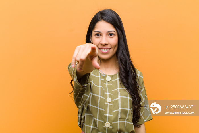 young pretty hispanic woman pointing at camera with a satisfied, confident, friendly smile, choosing you against brown wall