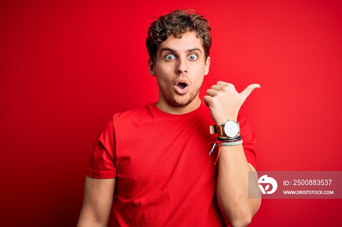 Young blond handsome man with curly hair wearing casual t-shirt over red background Surprised pointing with hand finger to the side, open mouth amazed expression.