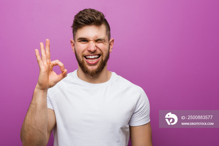 Young handsome caucasian man winks an eye and holds an okay gesture with hand.