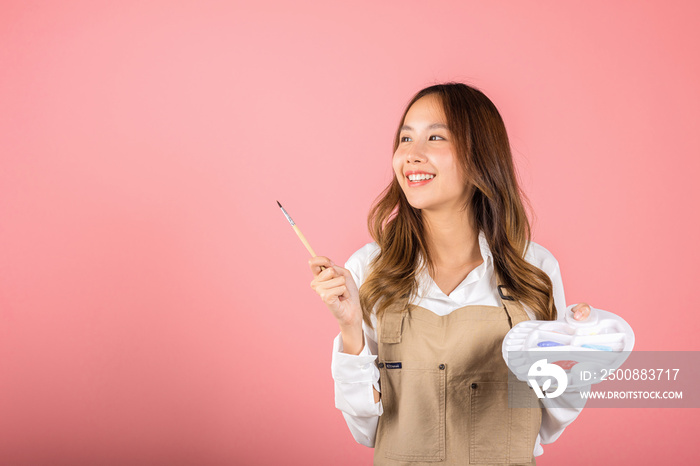 Happy female painting using paintbrush and palette with colors, Asian beautiful young woman artist holding brush and paint palette, studio shot isolated on pink background, Paintings and art equipment