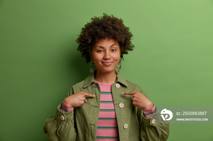 This is me! Satisfied proud Afro American woman points at herself, stands self confident, dressed fashionably, feels successful of her own achievement, stands indoor against green background