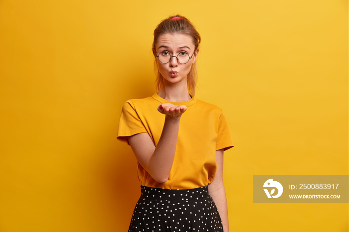 Beautiful female model with pony tail keeps lips folded holds palm forward near mouth blows air kiss expresses love to someone dressed in casual t shirt and skirt isolated on yellow background