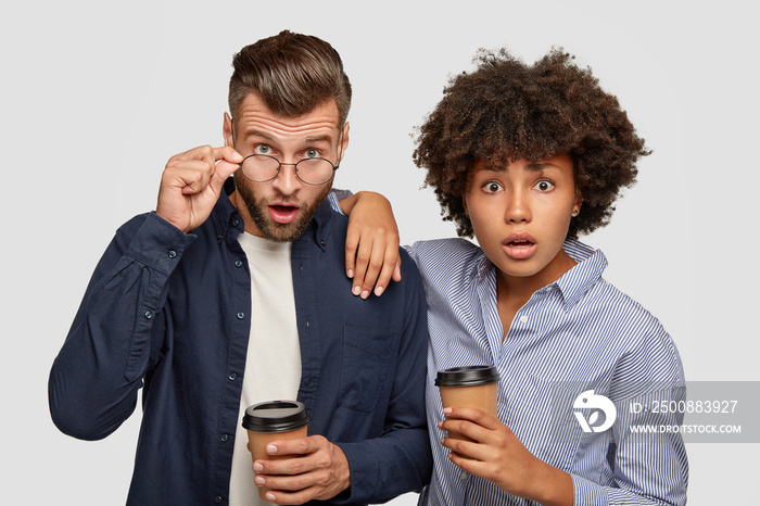 Picture of astonished mixed race woman and man stare with surprised epressions at camera, cannot believe own eyes, drink coffee, pose against white background together. Interracial couple indoor