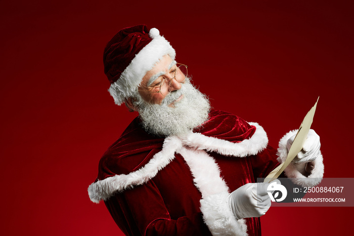 Waist up portrait of smiling Santa reading list on parchment standing against red background, copy space