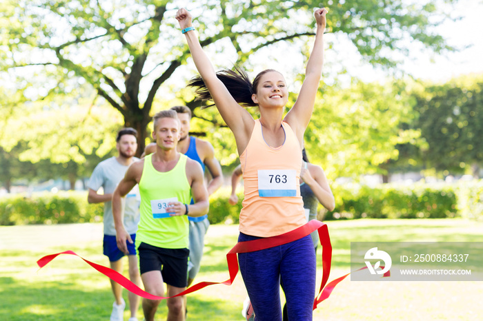 fitness, sport and success concept - happy woman winning race and coming first to finish red ribbon over group of sportsmen running marathon with badge numbers at park