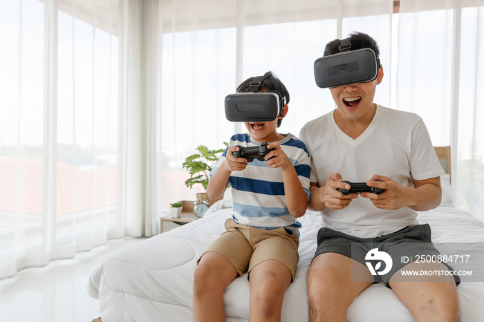 A father and son Sitting in bed. Asian family have fun and enjoy playing the game by wearing virtual glasses. On the bed in the room. Holiday concept..