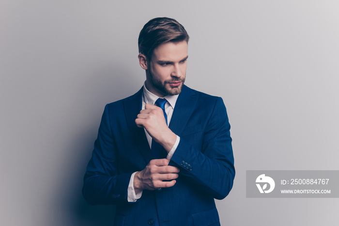 Portrait of stunning, fashionable, cool, rich man with stubble in blue suit correcting button on cuffs of sleeve of white shirt with arm, looking to the side, isolated on grey background