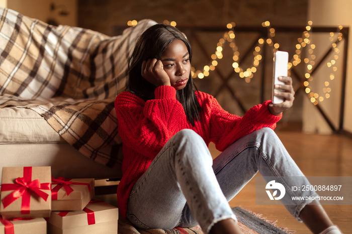 Unhappy young black woman looking at smartphone screen, sitting on floor near Christmas gift boxes at home
