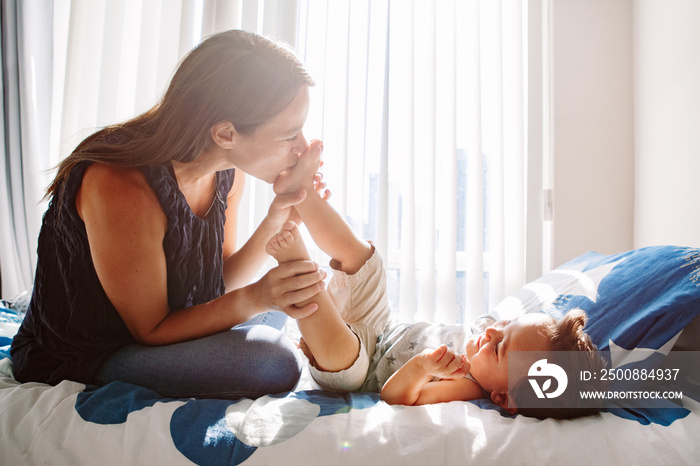 Young Caucasian mother kissing boy toddler son feet heels legs. Mom playing with child baby on bed in bedroom at home. Happy authentic family childhood lifestyle. Tender touching sweet moments.