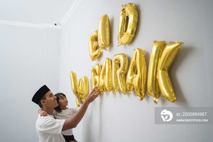 portrait father and daughter muslim decorating eid mubarak letter made of baloon decoration against the wall at home