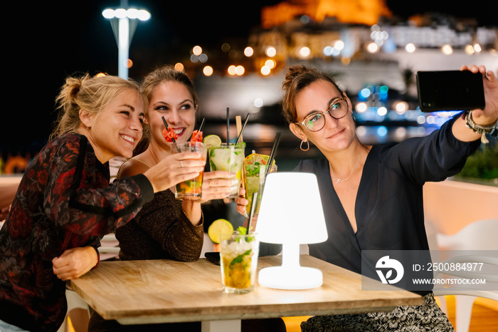 Three women making a selfie while sitting on a terrace drinking cocktails at night