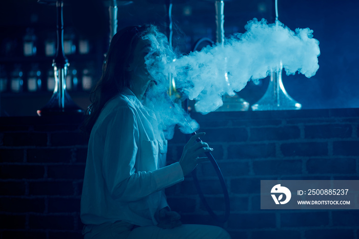 Beautiful woman blows smoke rings in night club hookah or shisha bar, blue background