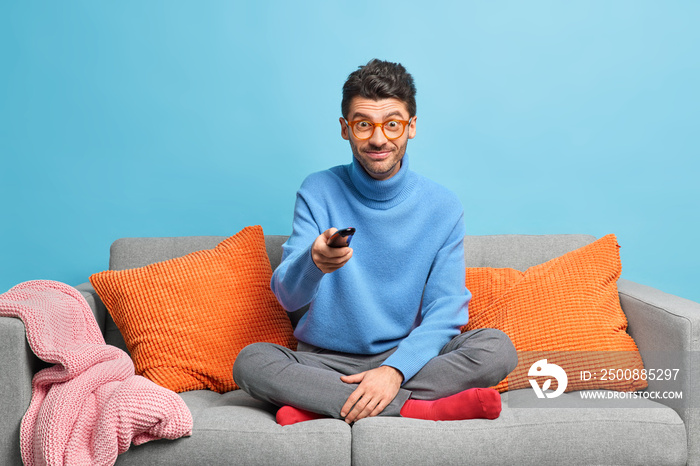 Pleased unshaven man changes TV channels with remote control watches funny program dressed in casual clothes sits crosses legs on comfortable sofa isolated over blue background. Pastime concept