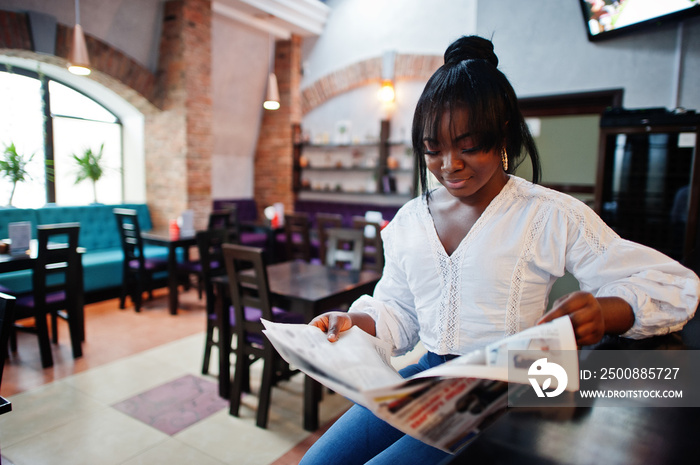 Stylish african american women in white blouse and blue jeans posed at cafe with newspaper.