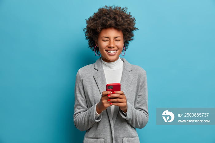 Cheerful Afro American female blogger posts images online, works via phone, laughs over funny video in internet, has fun and closes eyes, wears grey jacket, isolated on blue background, uses app