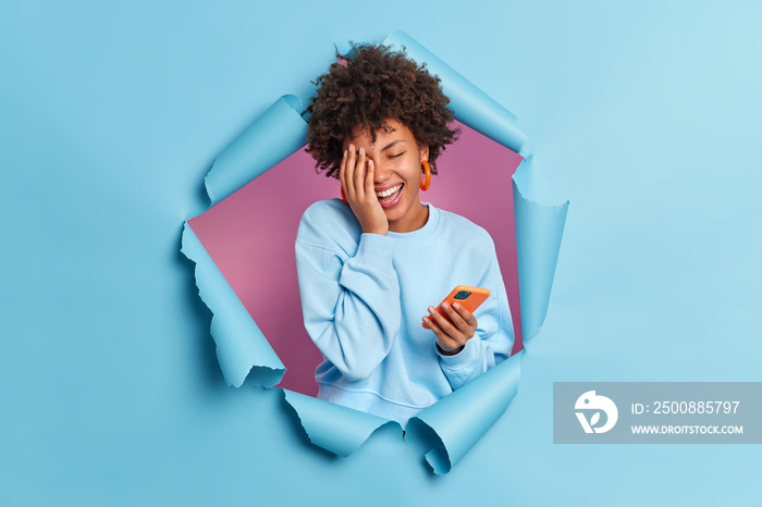 Photo of overjoyed young African American woman makes face palm smiles broadly keeps eyes closed uses smartphone for texting expresses positive authentic emotions breaks through blue paper wall