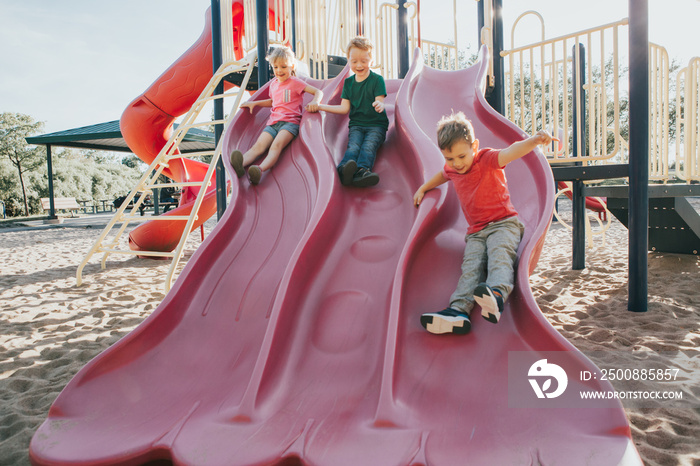 Active happy Caucasian children sliding on playground schoolyard outdoors on summer sunny day. Kid friends boys girl having fun. Seasonal kids activity outside. Authentic childhood lifestyle concept.