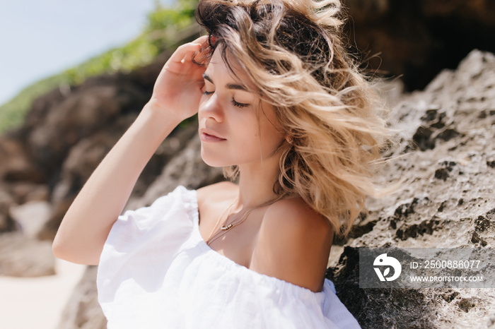 Pleasant girl posing near rock with eyes closed. Outdoor portrait of tanned sensual lady standing on nature background.