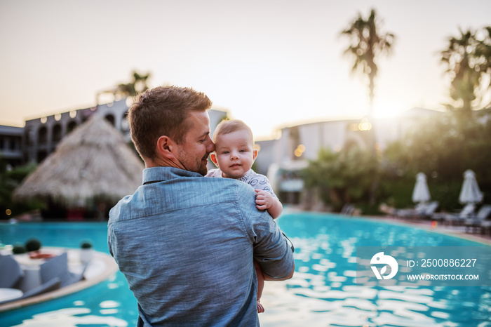 Handsome Caucasian dad hugging son and looking at him while standing next to swimming pool. Baby looking over father’s shoulder.