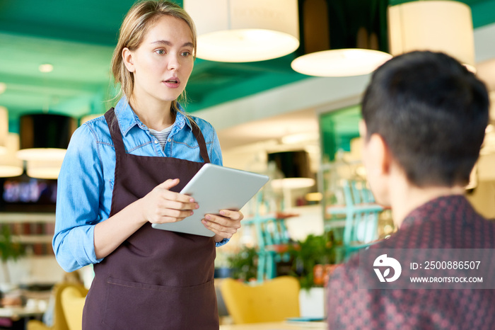 Portrait of young cafe waitress  wearing apron taking order from client using digital tablet, copy space