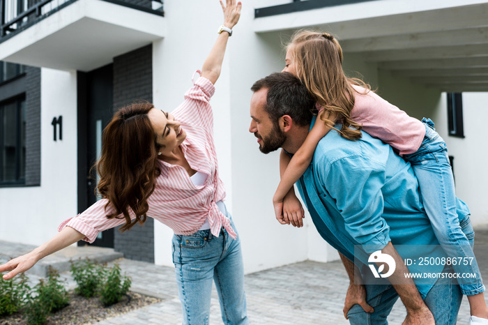 happy man piggybacking daughter near cheerful wife with outstretched hands