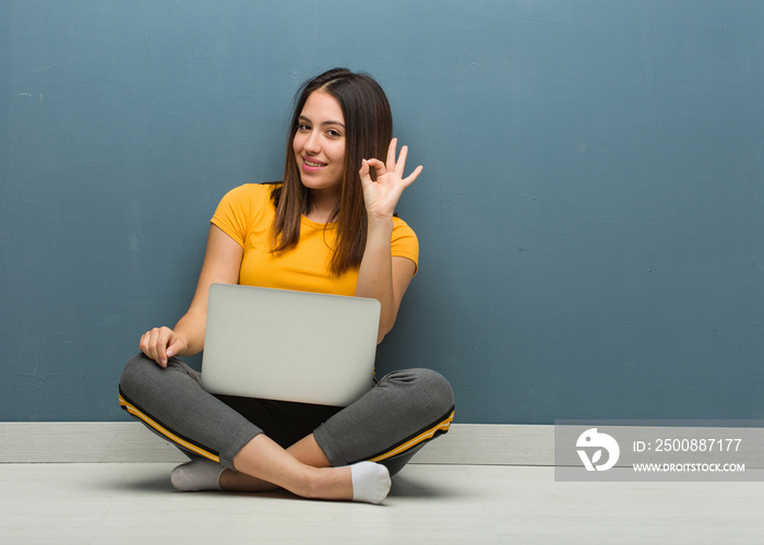 Young woman sitting on the floor with a laptop cheerful and confident doing ok gesture