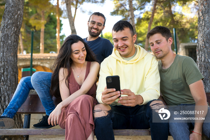 Group of friends sitting on a park bench. They are smiling while looking at their mobile phones. They are happy and wearing summer clothes. In a park with pine trees. Caucasian. Javea, Alicante.