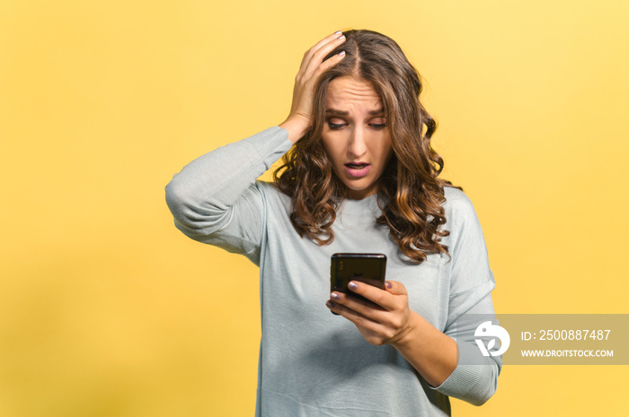 Shocked curly brunette young woman holding smartphone, looking at mobile phone screen and has disappointment face, received a bad news, made a mistake and a phone is locked, isolated on yellow