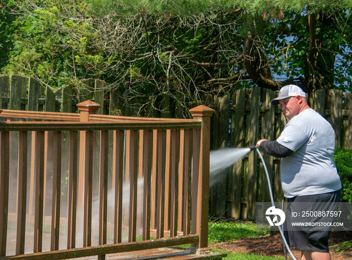 Overweight man wearing white baseball cap power washing the railing of a brown wooden deck in a backyard