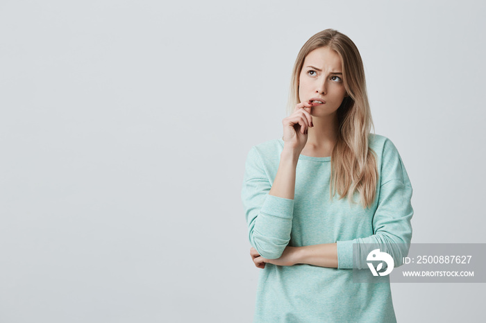 Beautiful blonde female with puzzled expression, keeps finger on lips, looks aside with bewilderment, poses against gray background. Body language and face expression