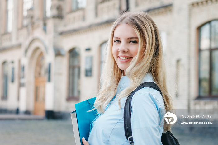 A blonde student girl is smiling and holding a folder and a notebook in her hands on a university background. Girl is taking exams at university