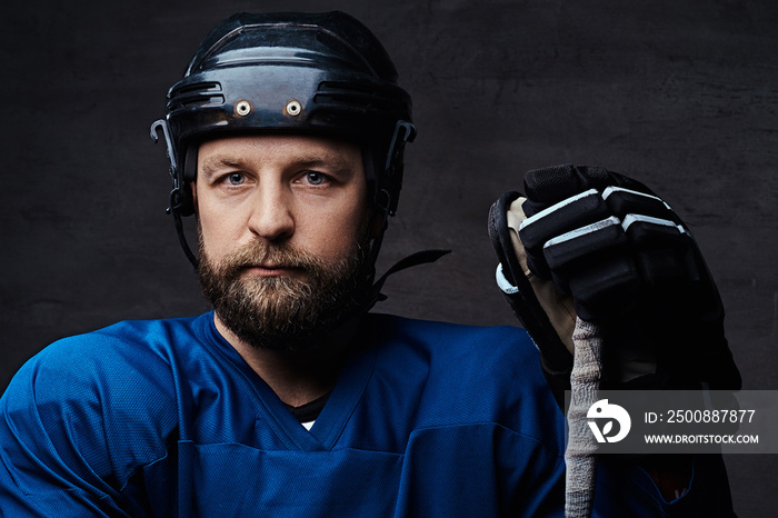 Close-up portrait of a middle-age bearded hockey player wearing full sports equipment with a hockey stick, over a dark background.