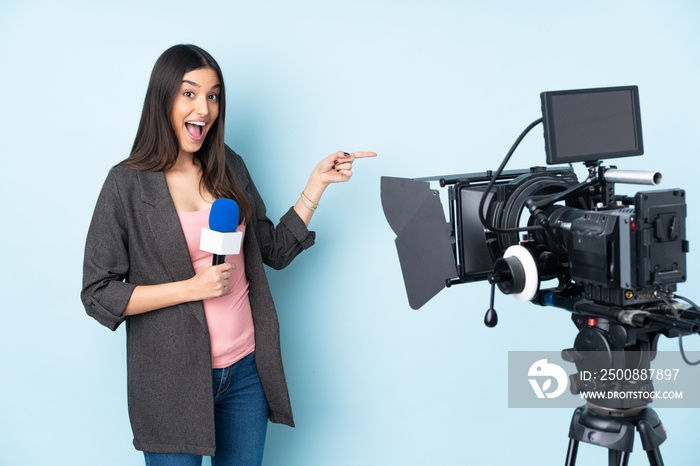 Reporter woman holding a microphone and reporting news isolated on blue background surprised and pointing side