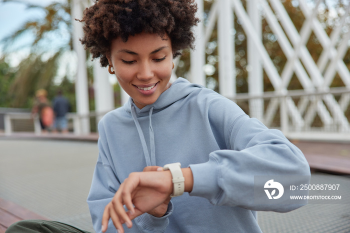 Attractive positive young Afro American woman checks time on wrist watch glad not to be late on meeting wears casual hoodie sits outdoors against blurred background feels relaxed and confident