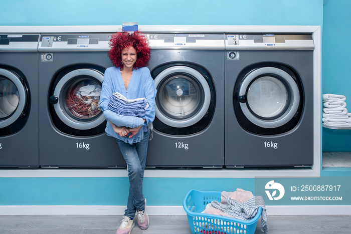 Smiling young woman with red afro hair holding clothes in a blue automatic laundromat