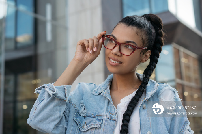 Portrait of beautiful African American woman wearing stylish eyeglasses, standing on the street and smiling. Young happy fashion model posing for pictures outdoors. Natural beauty concept