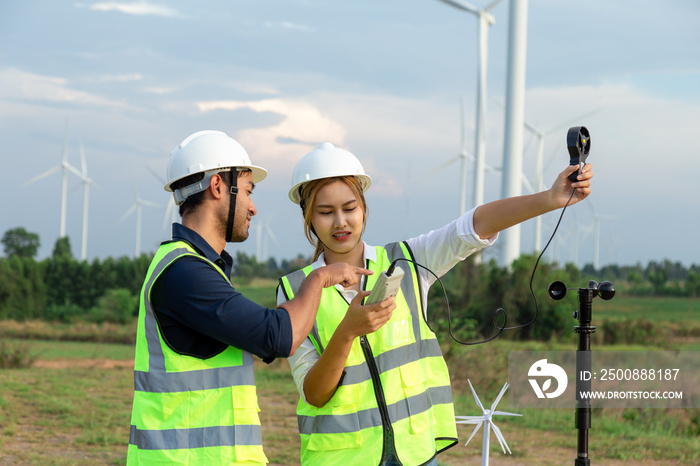 Two young engineers use an anemometer to measure wind speed, temperature and humidity on a wind farm background.