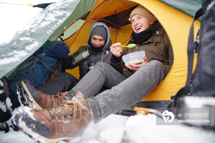 The guys are sitting in a tent, they are preparing food during the winter trip. Near the tent in the snow is their equipment