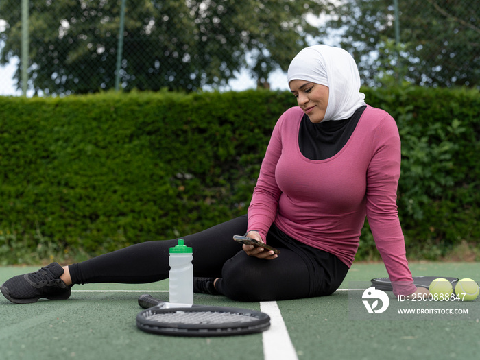 UK,Sutton,Woman in headscarf sitting on tennis court,looking at smart phone