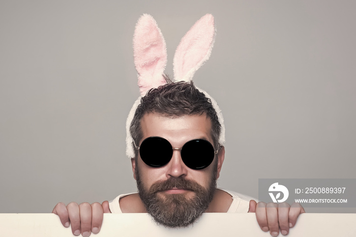 Funny man with beard in bunny ears and glasses on serious face with paper on grey background