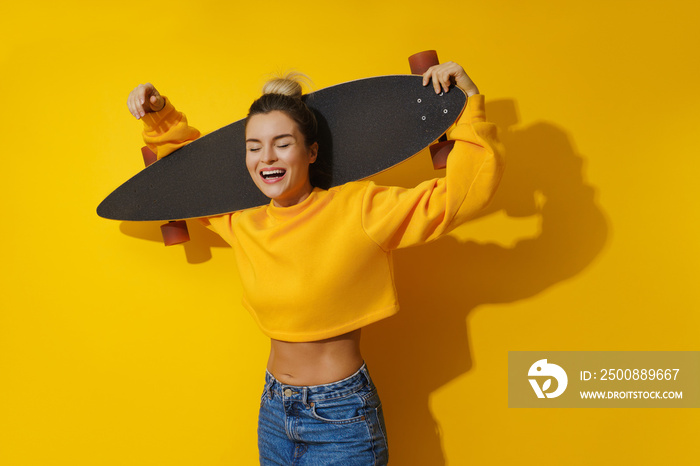 Young cheerful girl with longboard against yellow background